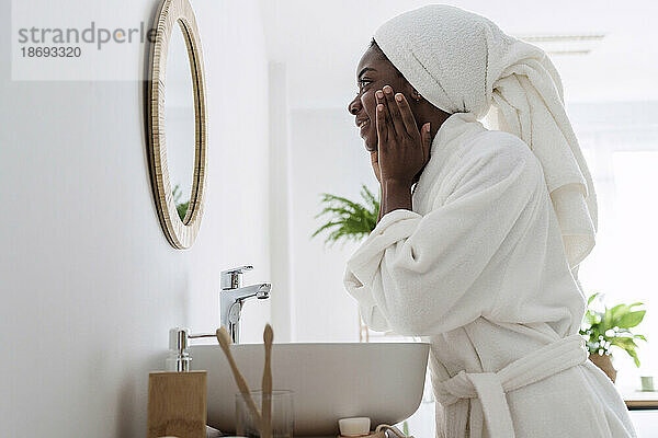 Frau trägt Bademantel und trägt zu Hause im Badezimmer Feuchtigkeitscreme auf ihr Gesicht auf