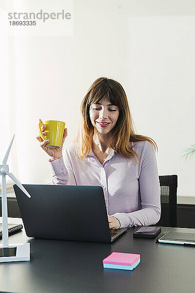 Lächelnde Geschäftsfrau hält Kaffeetasse in der Hand und arbeitet am Laptop im Büro