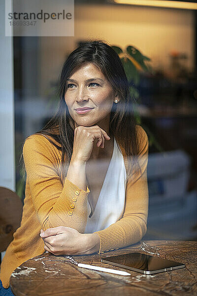 Frau mit Hand am Kinn sitzt im Café und sieht durch Glasfenster