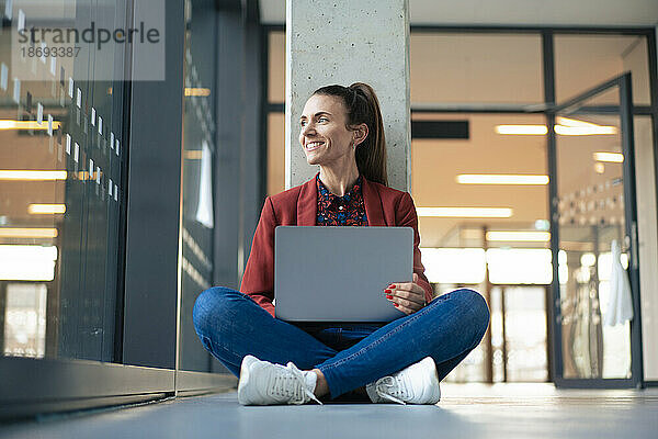 Lächelnde Geschäftsfrau mit Laptop sitzt im Schneidersitz auf dem Boden im Büro