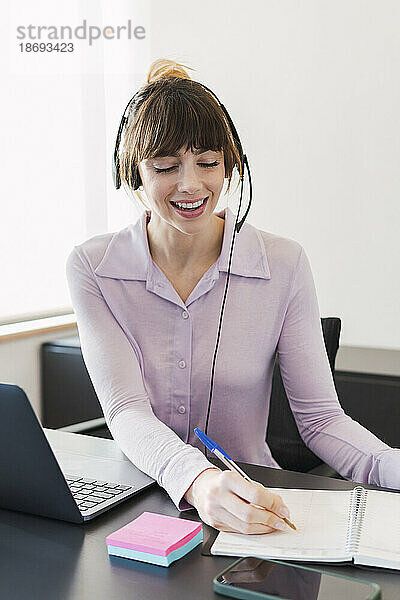 Glückliche Geschäftsfrau mit Headset schreibt auf Notizblock am Schreibtisch