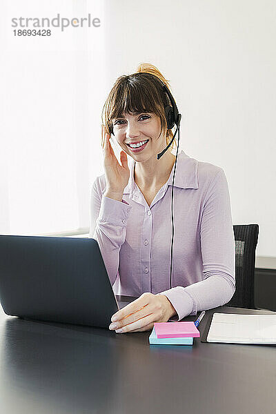 Glückliche Geschäftsfrau mit Headset sitzt mit Laptop am Schreibtisch