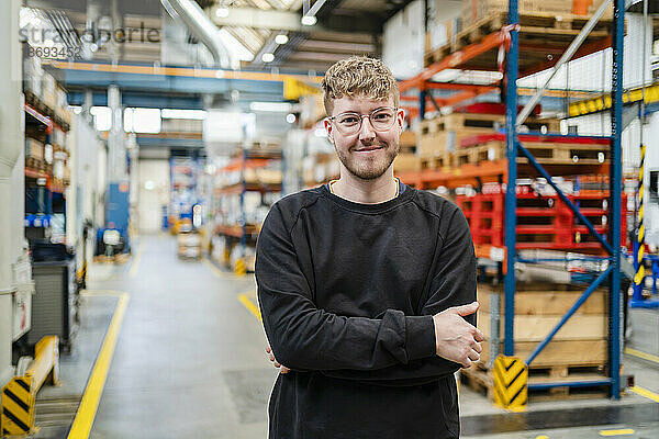 Lächelnder junger Mitarbeiter mit verschränkten Armen in der Fabrik