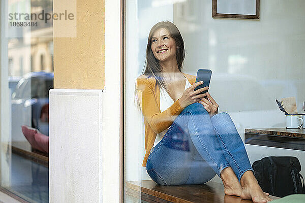 Lächelnde Frau mit Mobiltelefon sitzt im Café und sieht durch die Glasscheibe