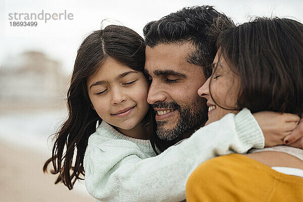 Lächelndes Mädchen mit geschlossenen Augen umarmt Mutter und Vater