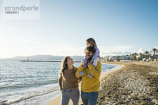 Vater trägt Tochter auf Schultern und spaziert mit Frau am Strand