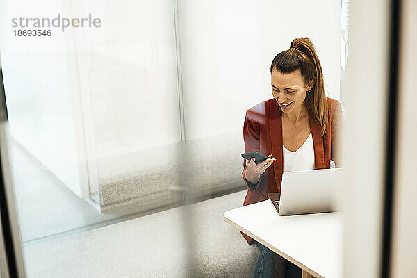Geschäftsfrau mit Laptop und Smartphone sitzt am Schreibtisch im Büro