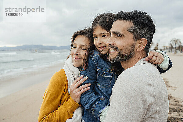 Lächelnde Frau mit Mann und Tochter  die sich am Strand umarmen
