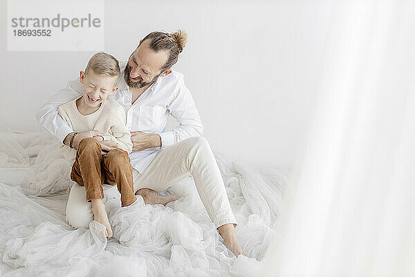 Man tickling son sitting near white wall