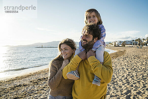 Lächelnde Tochter sitzt auf den Schultern des Vaters  neben einer Frau am Strand
