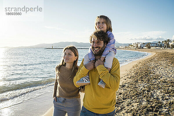 Lächelnde Eltern spazieren mit ihrer Tochter am Strand