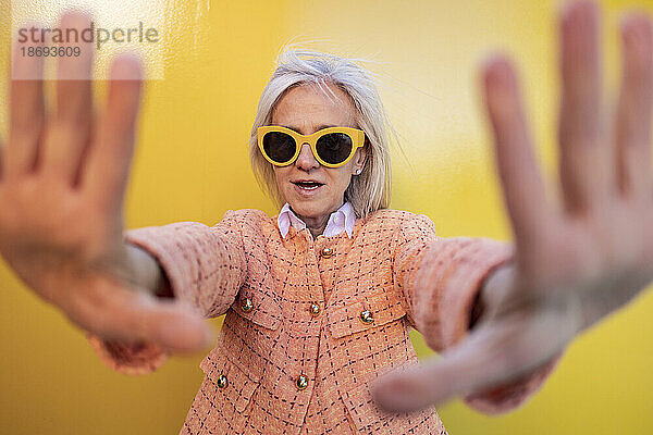Frau mit Sonnenbrille macht Stopp-Geste vor gelber Wand