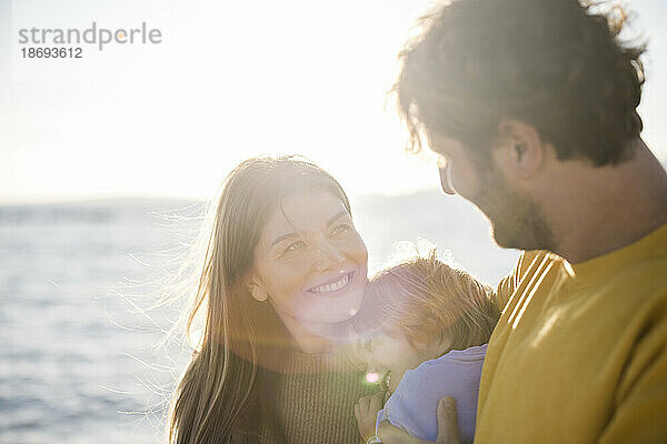 Lächelnde Frau blickt Mann an  der an einem sonnigen Tag seine Tochter am Strand hält