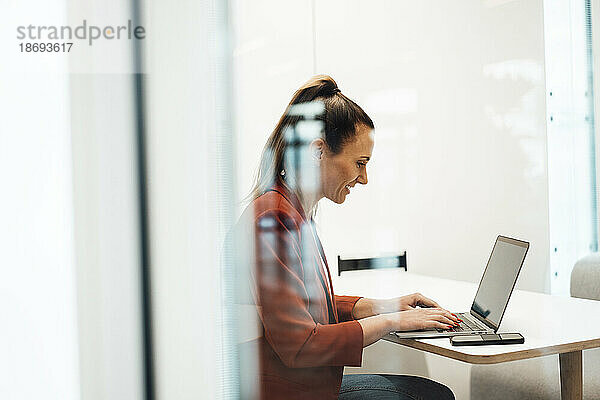 Geschäftsfrau  die am Laptop arbeitet und am Schreibtisch sitzt und durch eine Glaswand gesehen wird