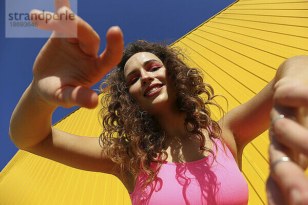 Lächelnde Frau gestikuliert an einem sonnigen Tag in der Nähe der gelben Wand