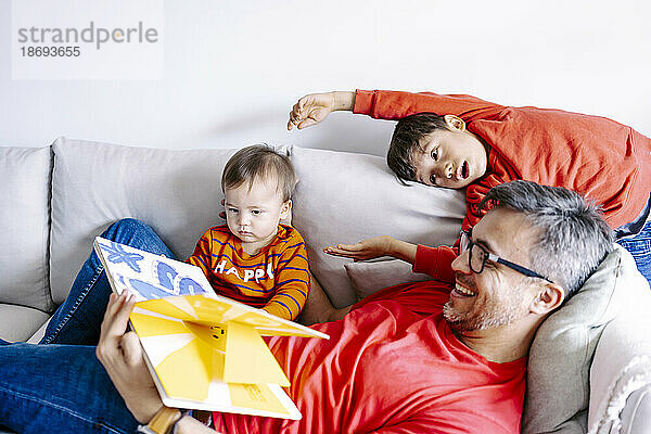 Vater zeigt seinen Söhnen zu Hause auf dem Sofa ein Buch