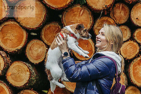 Lächelnde Frau hält Jack-Russell-Terrier-Hund vor Holzstämmen