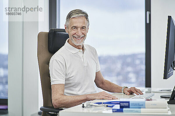 Porträt eines lächelnden Arztes  der in der Arztpraxis am Schreibtisch sitzt