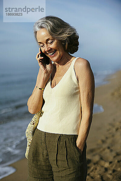 Glückliche Frau  die am Strand steht und mit ihrem Smartphone spricht