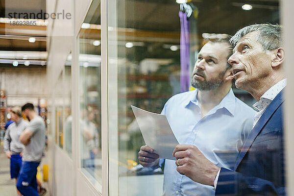Geschäftsmann und Manager diskutieren über ein Dokument  das durch ein Glasfenster in der Fabrik gesehen wird