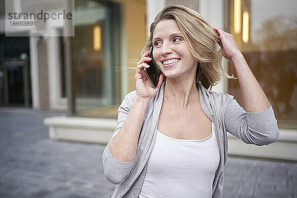 Junge lächelnde Frau mit der Hand im Haar  die auf dem Smartphone spricht