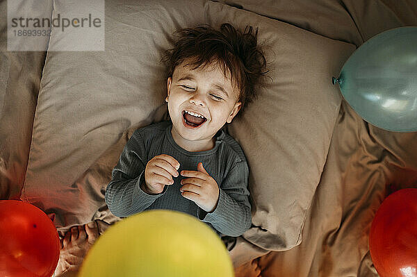 Boy laughing amidst balloons on bed