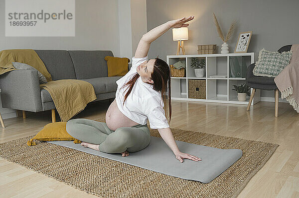 Schwangere Frau streckt die Hand  während sie zu Hause auf der Trainingsmatte sitzt