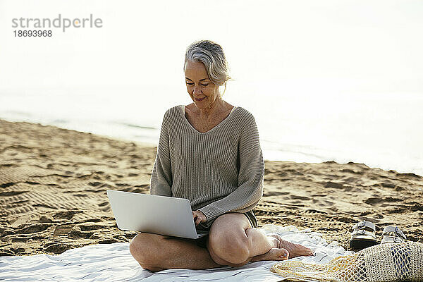 Reife Frau sitzt am Strand und benutzt Laptop