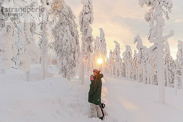 Frau mit Snowboard steht bei Sonnenaufgang auf einem schneebedeckten Feld