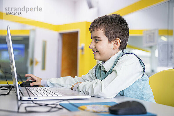 Lächelnder Junge  der in der Schule am Schreibtisch sitzt und einen Laptop benutzt
