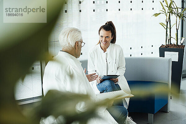 Lächelnder Arzt diskutiert mit einem älteren Kollegen  der auf der Couch im Krankenhaus sitzt