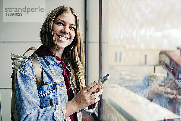Lächelnde junge Frau hält Smartphone am Glasfenster am Flughafen