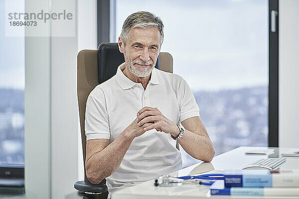 Porträt eines selbstbewussten Arztes  der in der Arztpraxis am Schreibtisch sitzt