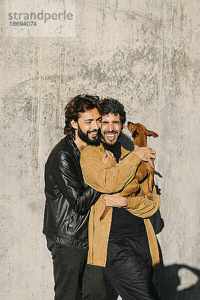 Happy gay couple enjoying with dog standing in front of concrete wall