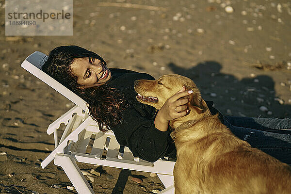 Lächelnde Frau liegt auf Liegestuhl und streichelt Hund am Strand