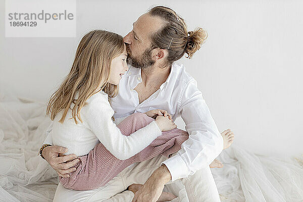Vater küsst Tochter  die neben weißer Wand sitzt
