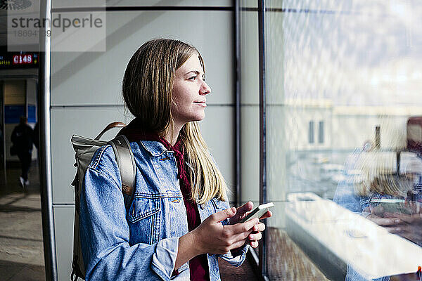 Junge Frau blickt mit Smartphone am Flughafen aus dem Fenster