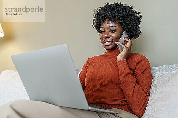 Lächelnde junge Geschäftsfrau benutzt Laptop und spricht zu Hause mit dem Smartphone