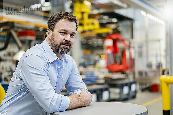 Lächelnder reifer Manager  der sich in der Fabrik auf den Tisch lehnt