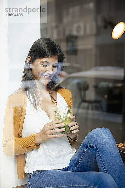 Lächelnde Frau hält gesundes Getränk in der Hand und sitzt im Café  gesehen durch die Glasscheibe