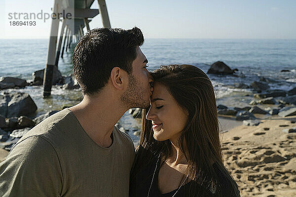 Junger Mann küsst Frau am Strand auf die Stirn