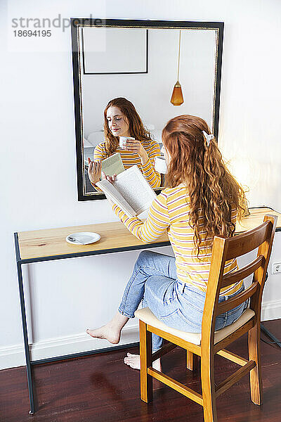 Frau mit Tasse in der Hand liest ein Buch und sitzt auf einem Stuhl vor dem Spiegel