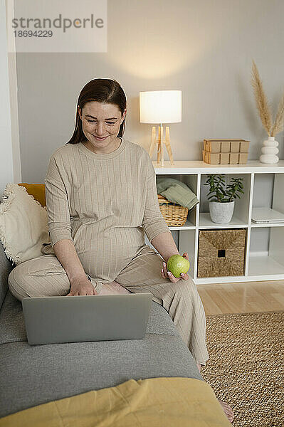 Lächelnde schwangere Freiberuflerin sitzt mit Laptop auf dem Sofa im Heimbüro