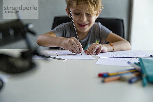 Lächelnder Junge  der am Schreibtisch mit Bleistift auf Papier zeichnet