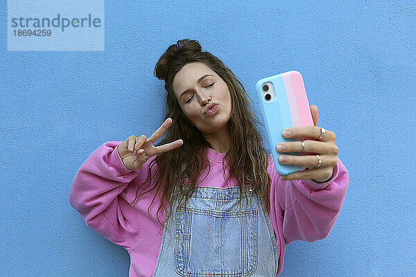 Young woman puckering and taking selfie with peace sign against blue background
