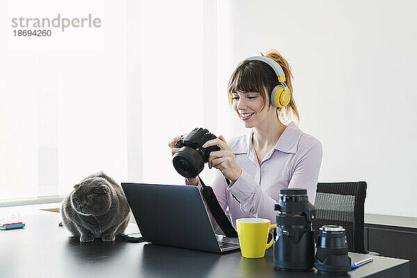 Glückliche Geschäftsfrau mit kabellosen Kopfhörern sitzt mit Kamera am Schreibtisch