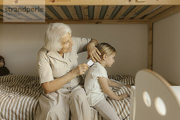 Großmutter kämmt die Haare ihrer Enkelin  die zu Hause im Bett sitzt