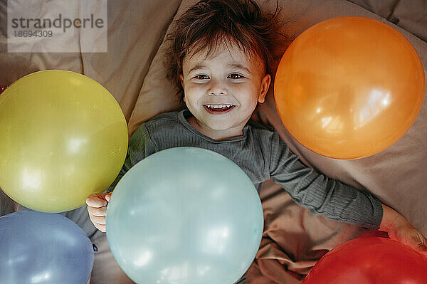 Glücklicher Junge  der inmitten von Luftballons auf dem Bett liegt