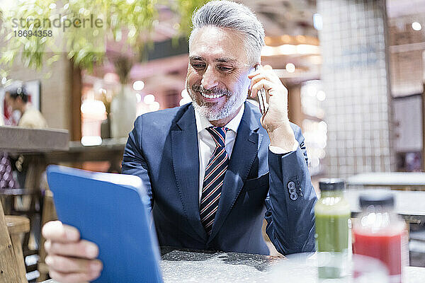 Lächelnder Geschäftsmann  der im Café mit einem Smartphone spricht und einen Tablet-PC in der Hand hält