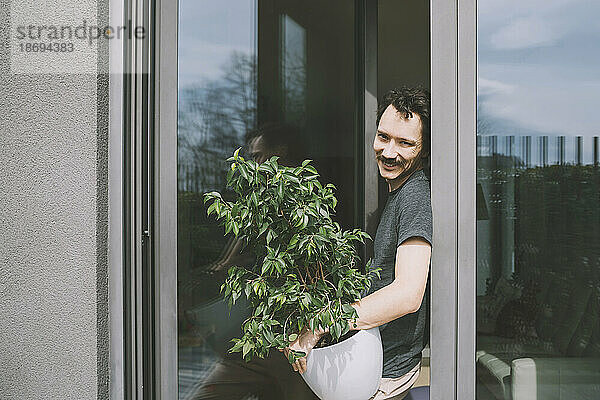 Happy man standing with potted plant by glass door